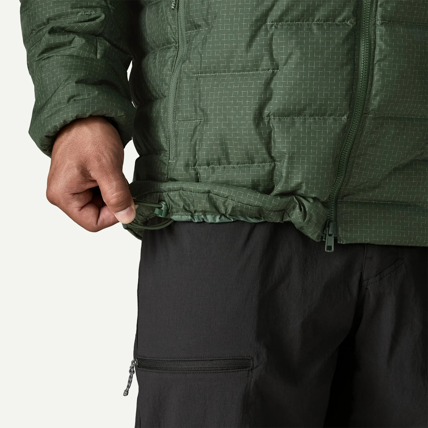 Close-up of a person wearing a dark green quilted puffer jacket and black pants, pulling the hem of the jacket down slightly with one hand against a light gray background.