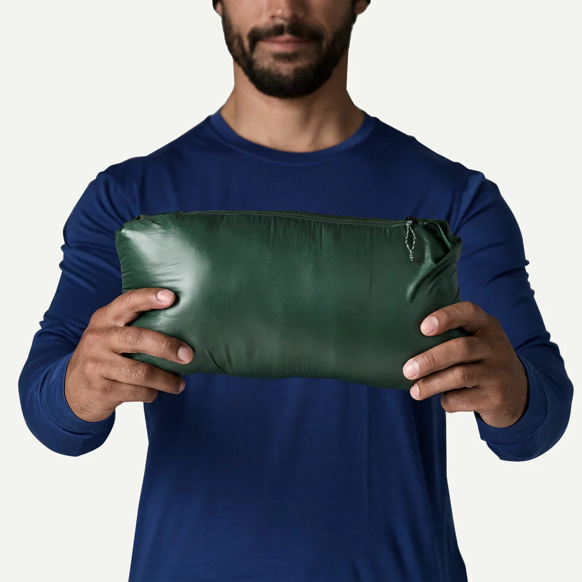 A man wearing a navy blue long-sleeve shirt holds a dark green, zippered pouch against a plain off-white background.