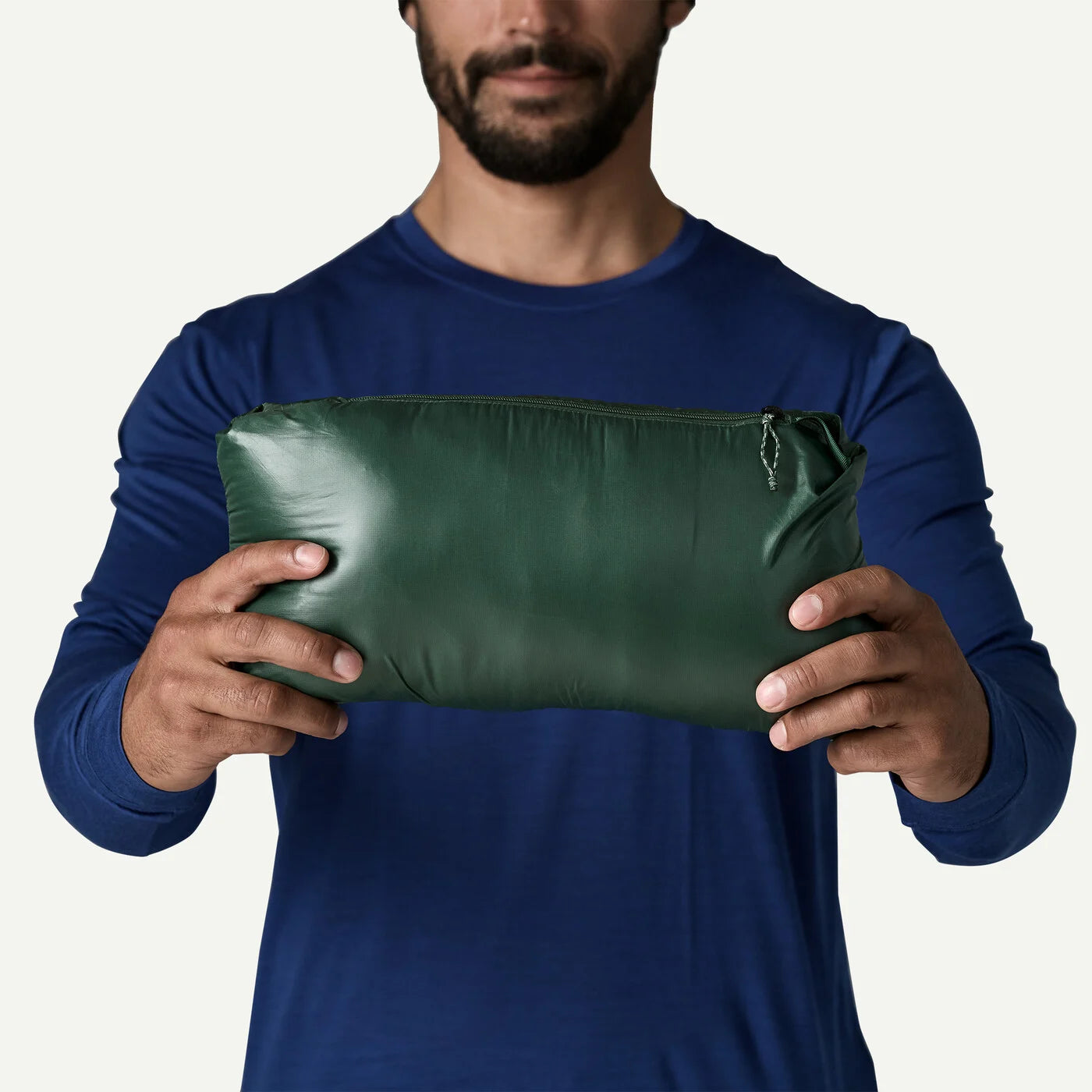 A man wearing a navy blue long-sleeve shirt holds a dark green, zippered pouch against a plain off-white background.