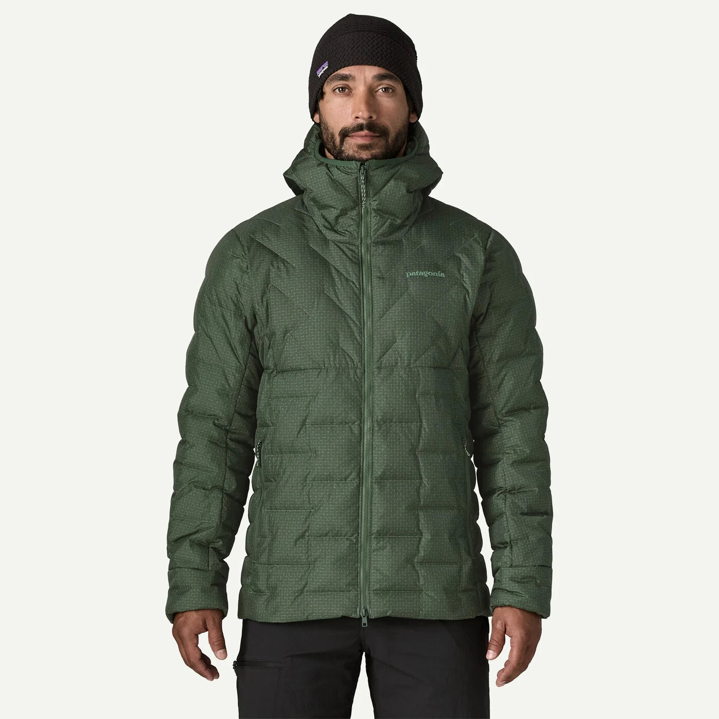 A man wearing a dark green quilted puffer jacket with a high collar, a black knit beanie, and black pants, standing against a solid off-white background.