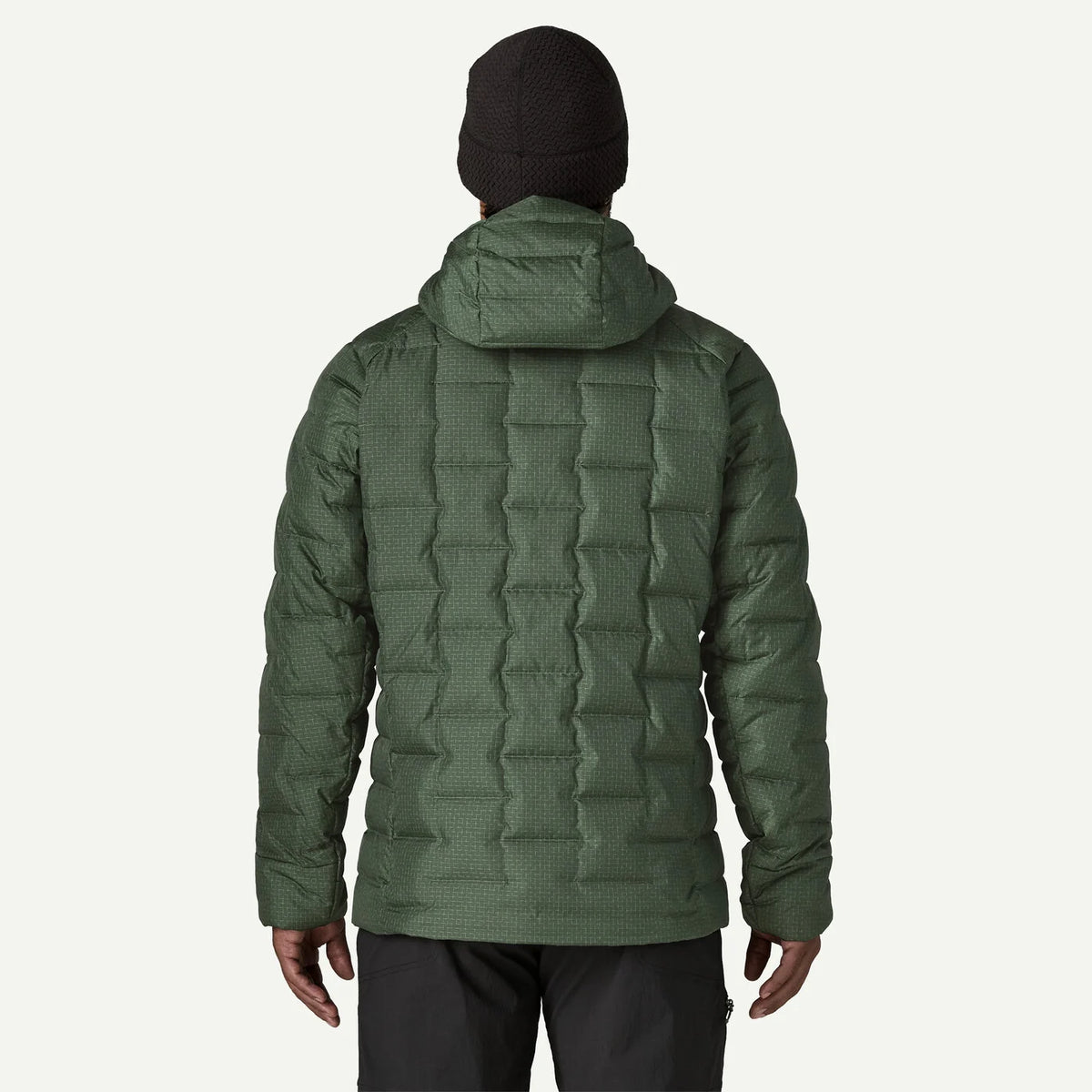 A man viewed from the back wearing an olive green quilted puffer jacket, a black beanie, and dark pants against a light gray studio background.