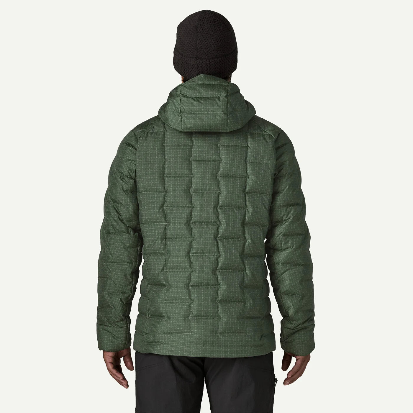 A man viewed from the back wearing an olive green quilted puffer jacket, a black beanie, and dark pants against a light gray studio background.