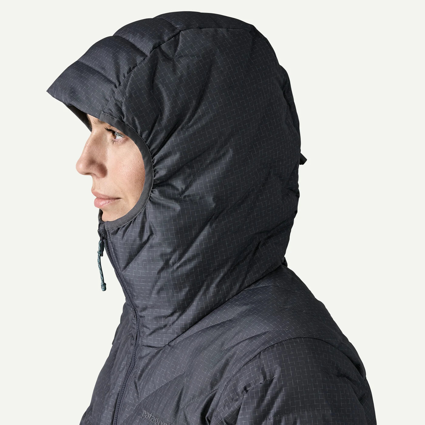 A close-up side profile of a person wearing a dark gray hooded puffer jacket against a light off-white background.