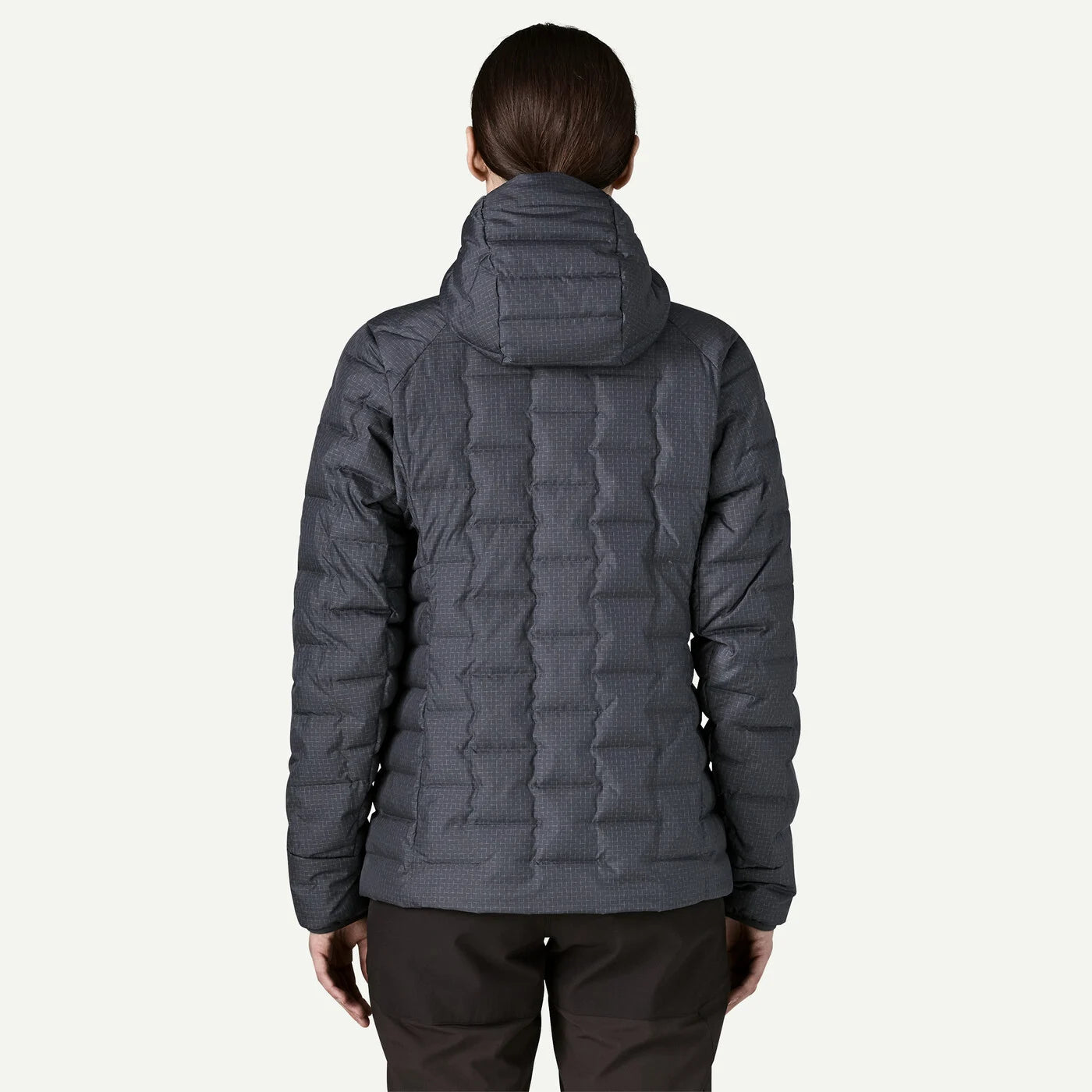 A woman viewed from the back wearing a dark gray quilted hooded puffer jacket and dark pants against a light gray studio background.
