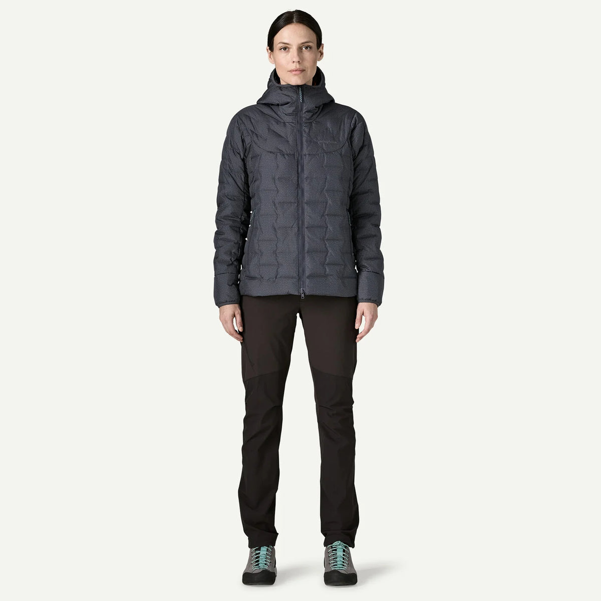 A woman modeling a dark gray hooded puffer jacket, paired with black pants and hiking shoes, standing against a solid off-white background.