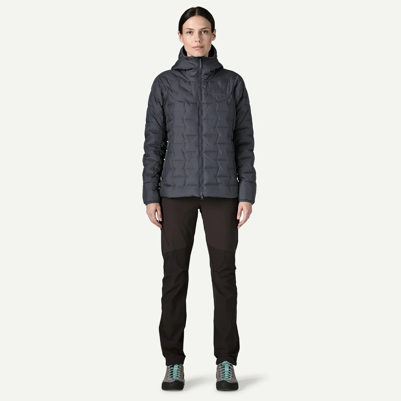 A woman modeling a dark gray hooded puffer jacket, paired with black pants and hiking shoes, standing against a solid off-white background.