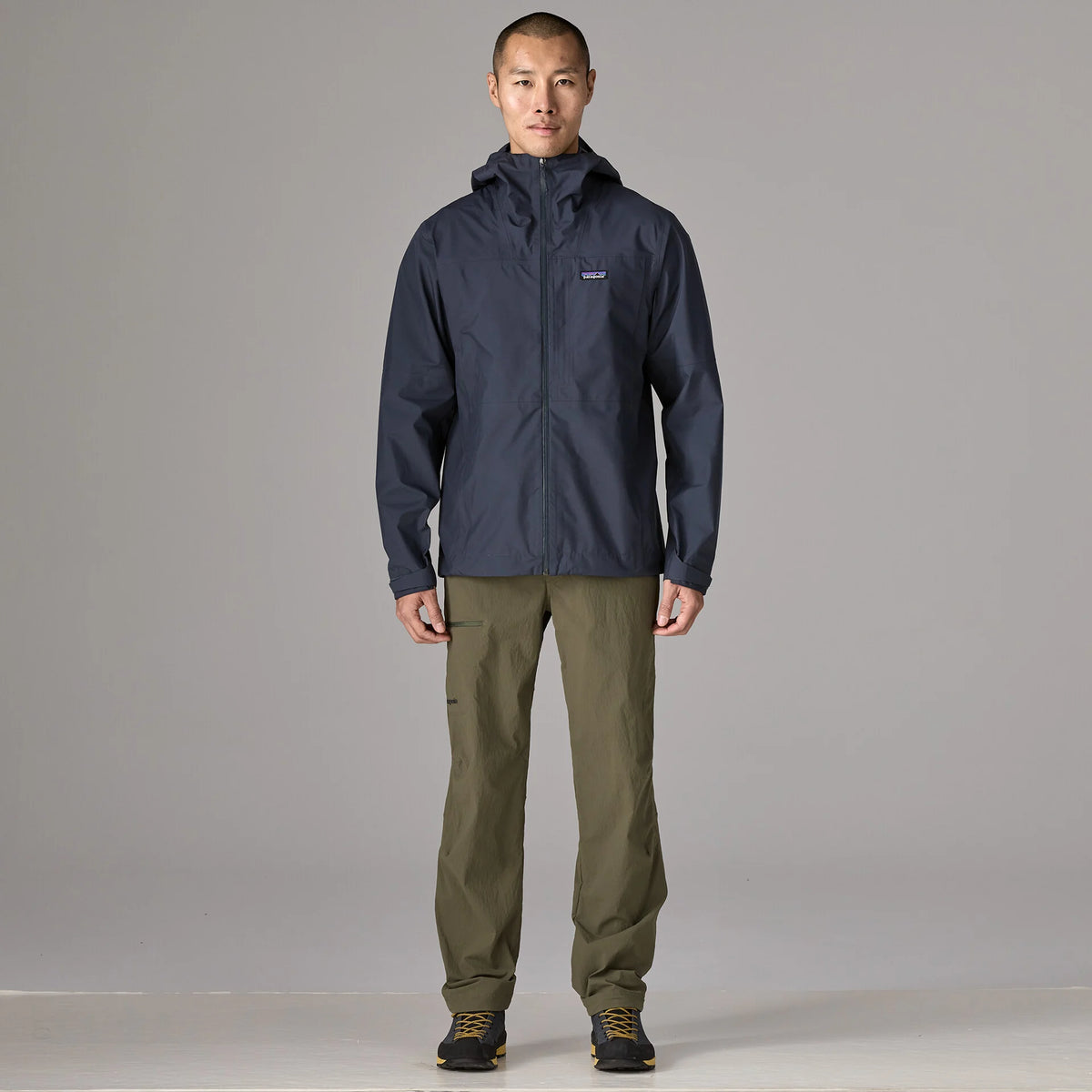 A man stands facing forward in a studio wearing a dark navy hooded jacket with a small Patagonia logo on the left chest, olive green pants, and dark shoes, against a light gray background.
