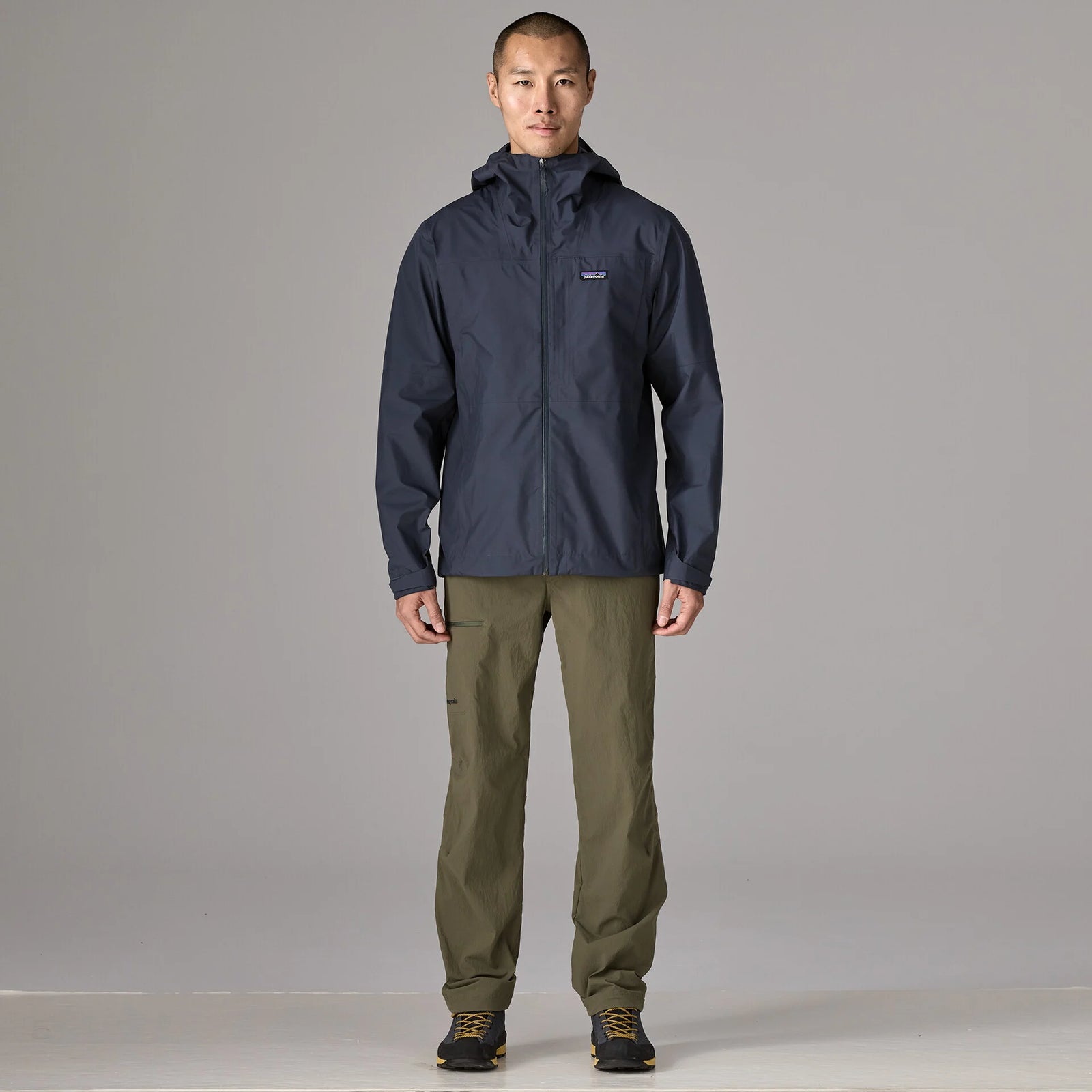 A man stands facing forward in a studio wearing a dark navy hooded jacket with a small Patagonia logo on the left chest, olive green pants, and dark shoes, against a light gray background.