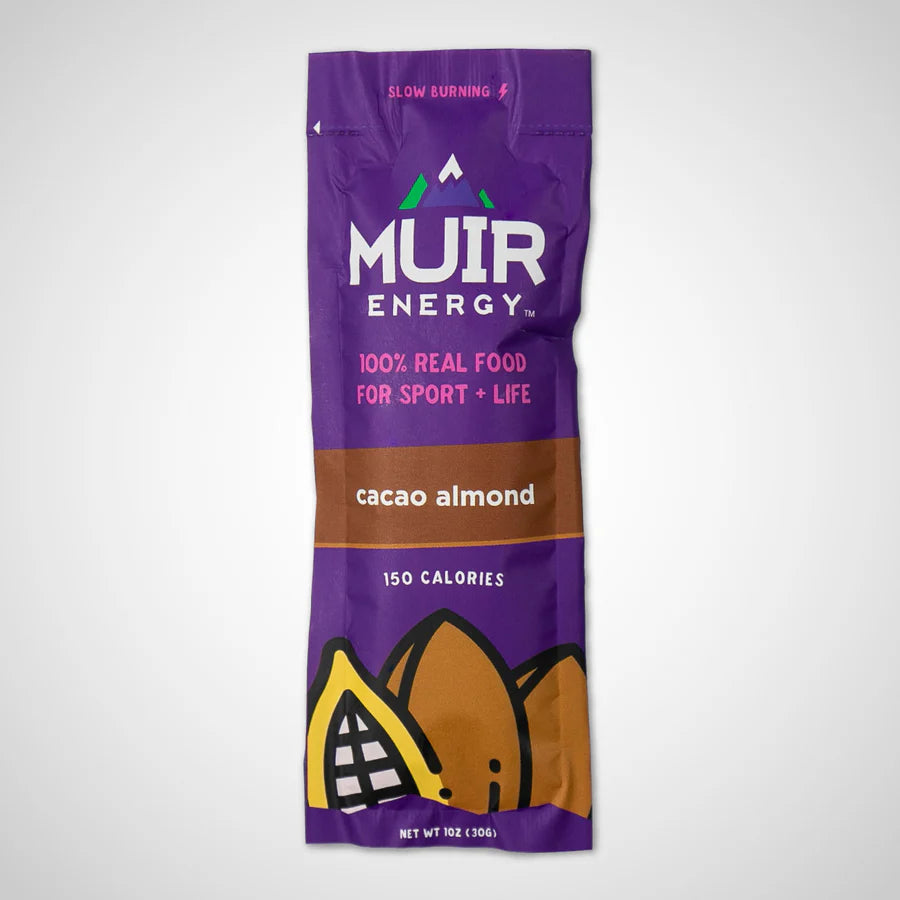 A single Muir Energy cacao almond snack stick pack on a light gray background. The packaging is primarily purple with a brown band in the middle and an illustration of almond slices at the bottom.