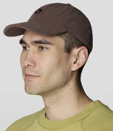 A man in profile wearing a brown corduroy baseball cap with a small black embroidered logo on the front, paired with an olive green sweatshirt, against a light gray background.