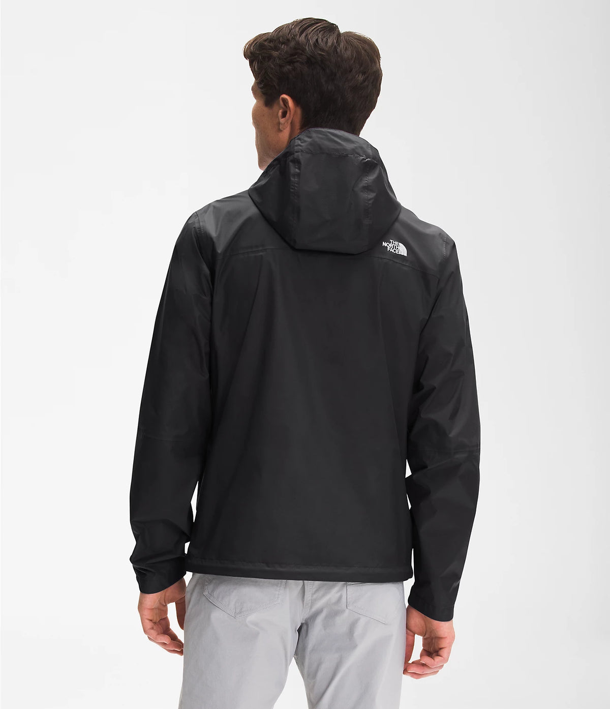 A man viewed from the back wearing a black hooded jacket with a small white The North Face logo on the upper back, paired with light gray pants, against a plain light gray studio background.