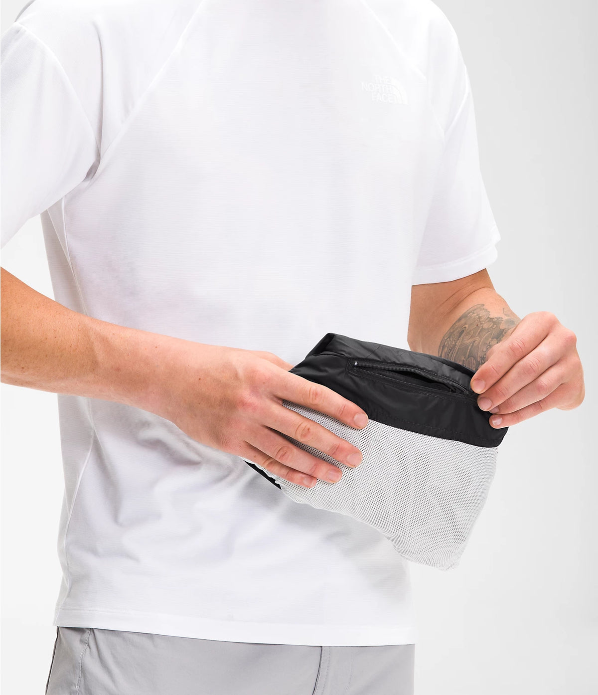A close-up of a person wearing a white t-shirt and light gray shorts, holding a black and light gray mesh waist bag or pouch in front of their torso.