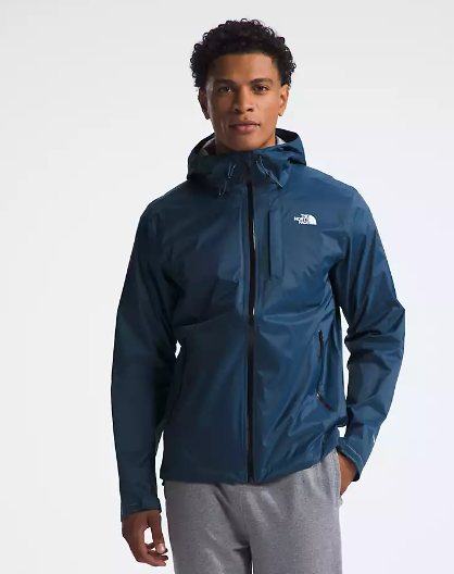 A man wearing a dark blue hooded rain jacket with a white embroidered logo on the left chest, paired with gray sweatpants, photographed against a light gray studio background.