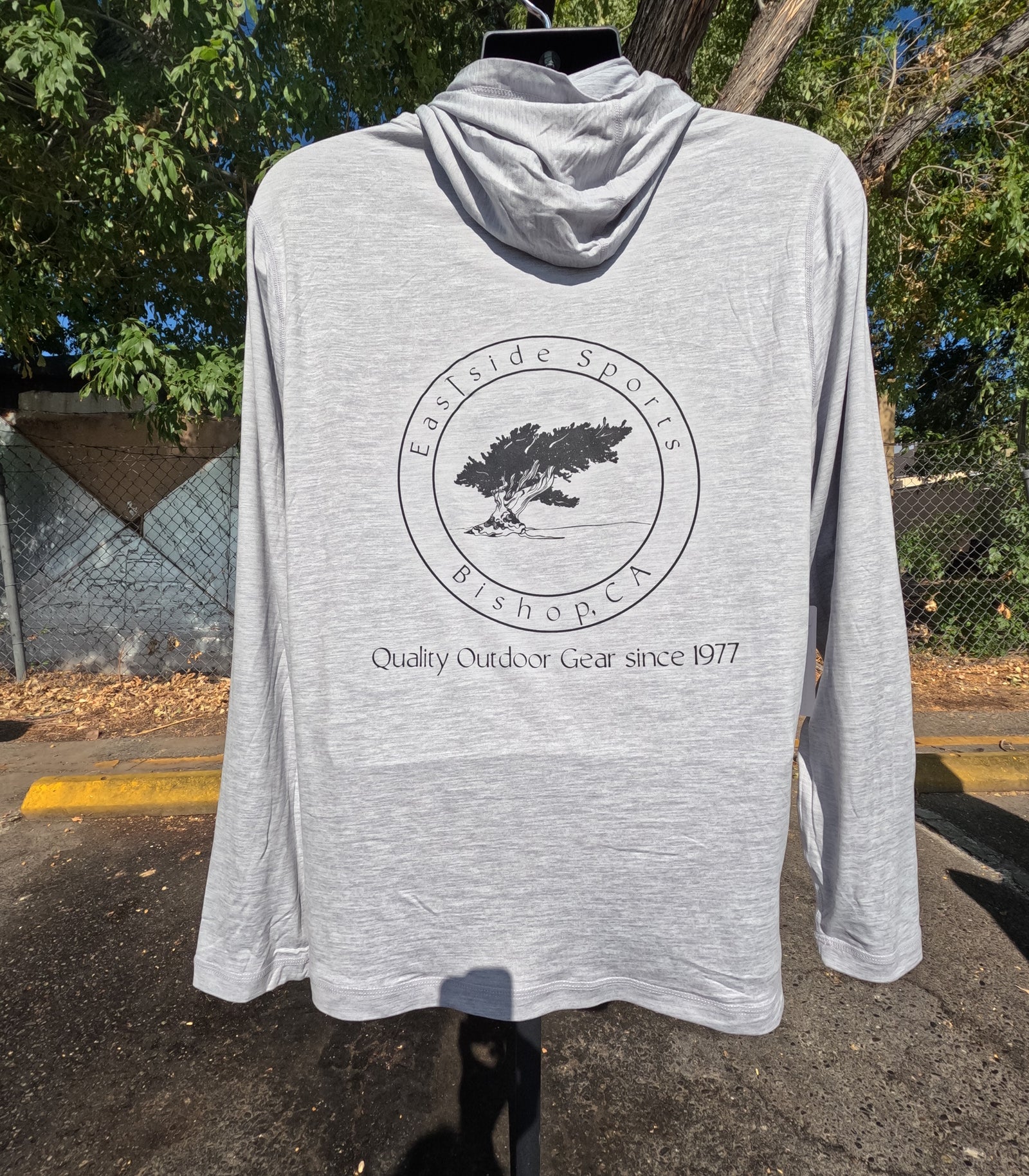 A light heather gray hooded long-sleeve shirt displayed on a hanger outdoors. The back of the shirt features a black circular graphic with a tree and the text 'Eastside Sports Bishop, CA' and 'Quality Outdoor Gear since 1977'.