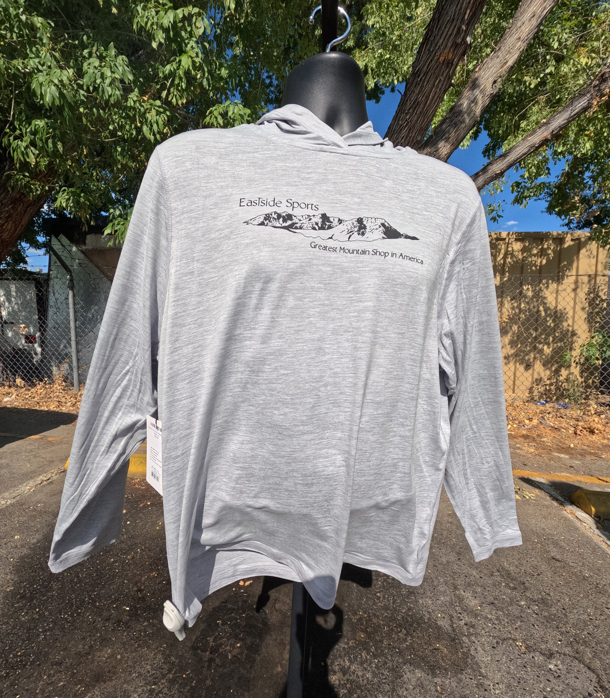 A light heather gray long-sleeve hooded shirt displayed on a black mannequin outdoors. The front has a black mountain graphic with the text 'Eastside Sports' and 'Greatest Mountain Shop in America.