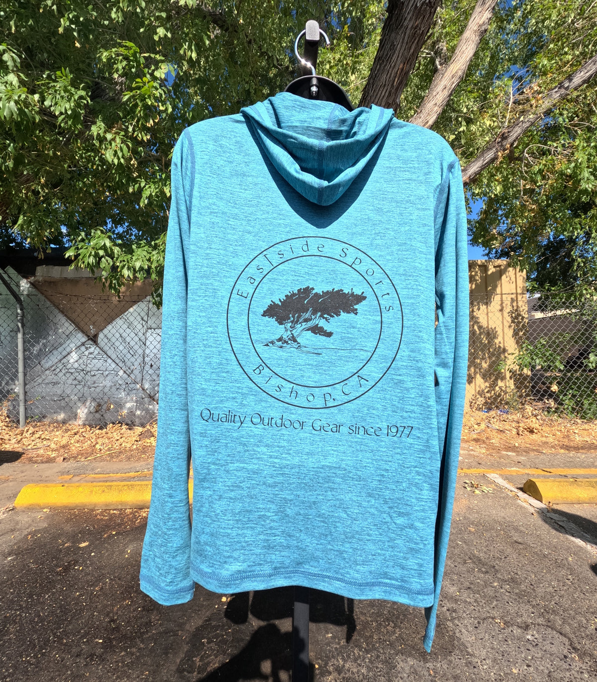 A heathered blue long-sleeve hooded shirt hanging on a black hanger outdoors. The back of the shirt features a circular black graphic with a tree and the text 'Eastside Sports Bishop, CA' and 'Quality Outdoor Gear since 1977'.