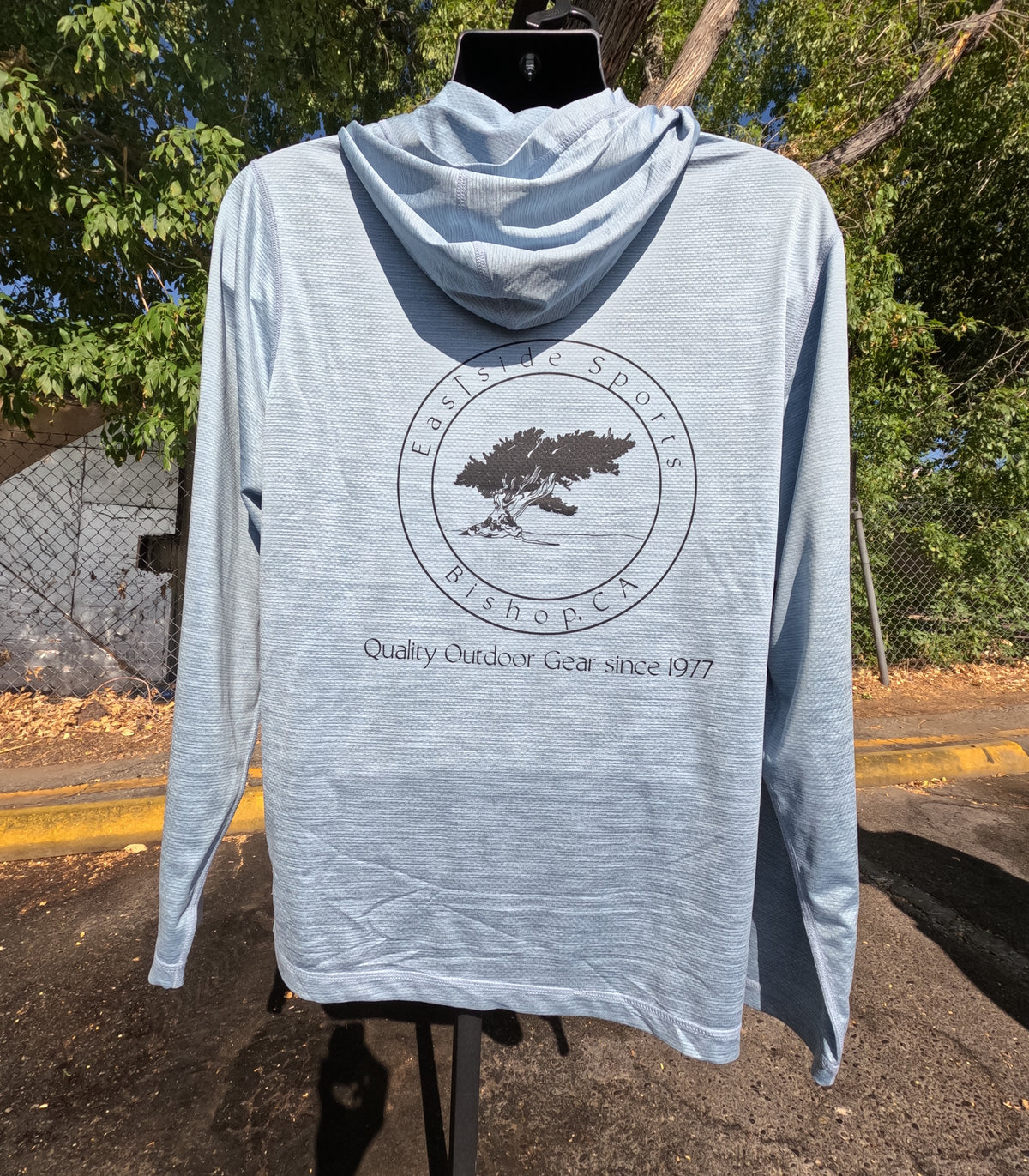 A light heather gray hooded long-sleeve shirt displayed on a black hanger outdoors. The back of the shirt features a circular black graphic with a tree and the text 'Eastside Sports Bishop CA' and 'Quality Outdoor Gear since 1977'.