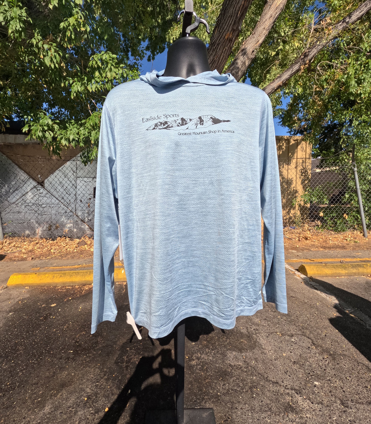 A light blue long-sleeve hooded shirt displayed on a black mannequin outdoors. The shirt features a small black graphic of mountains and text on the chest reading 'Eastside Sports' and 'Greatest Mountain Shop in America'.