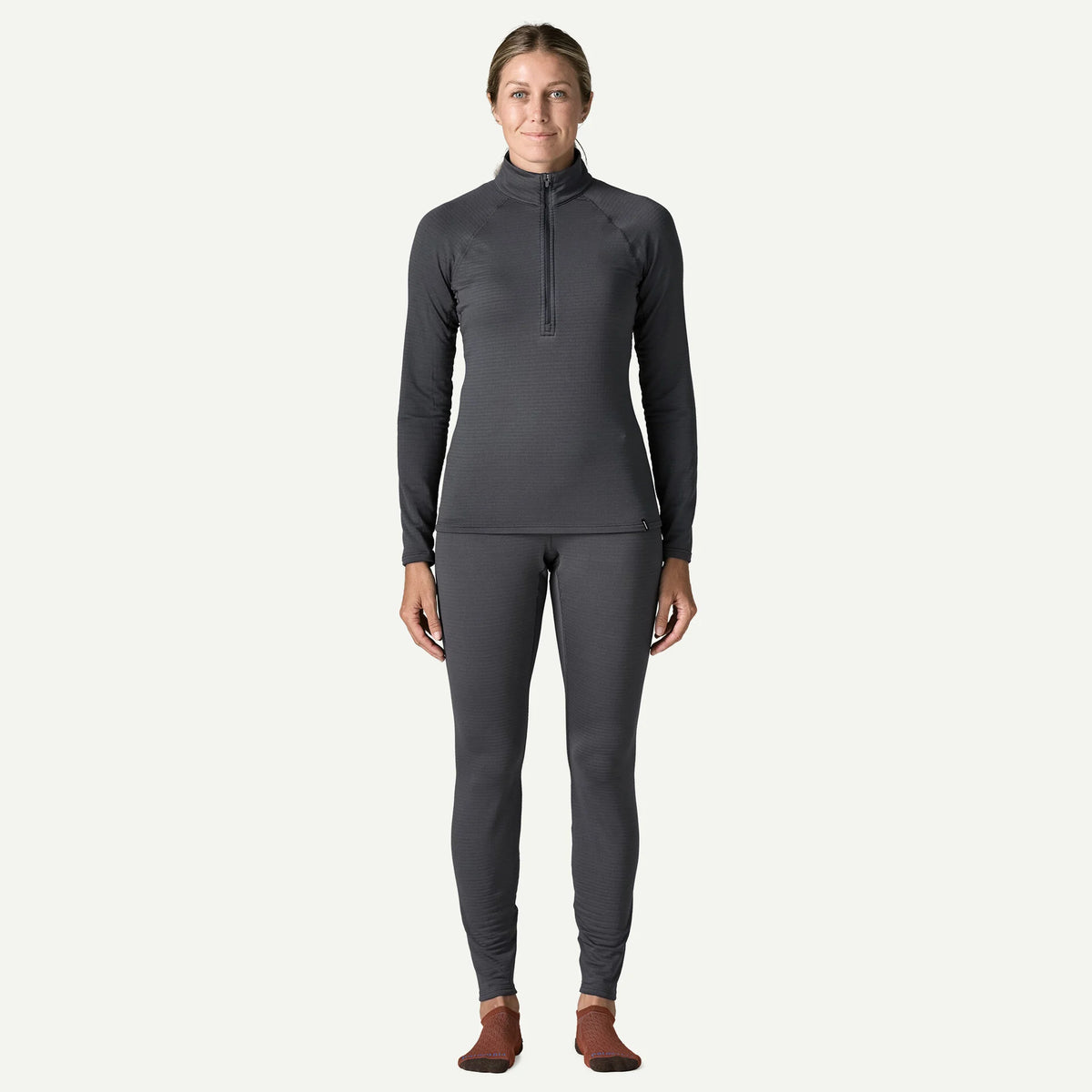 A woman modeling a dark gray long-sleeve quarter-zip top and matching full-length leggings against a light gray studio background.
