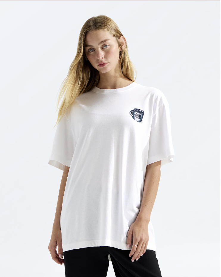 A woman modeling a white short-sleeve t-shirt with a small black and white graphic on the left chest, paired with black pants, against a light gray studio background.