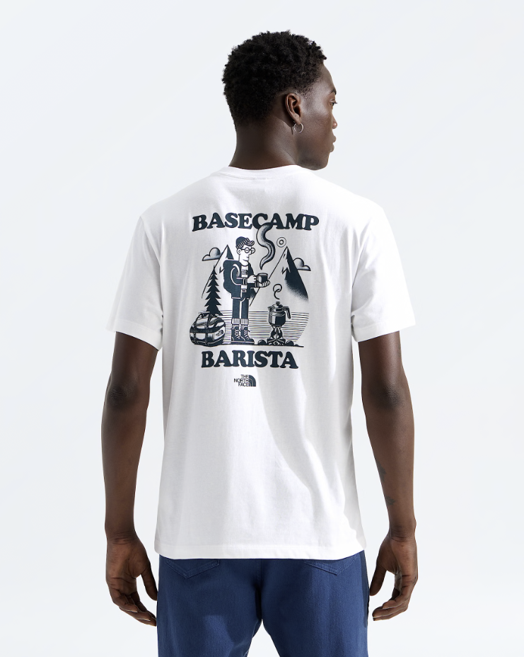 A man viewed from the back wearing a white short-sleeve t-shirt with a large navy blue graphic print on the back that reads 'BASECAMP BARISTA' with an illustration, and navy blue shorts, against a light gray studio background.