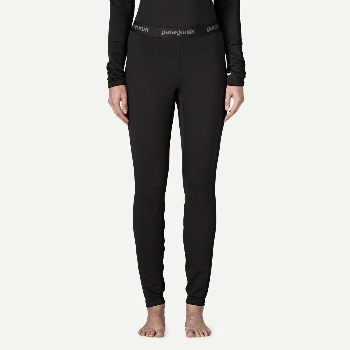 A model wearing black Patagonia full-length leggings and a black long-sleeve top, standing barefoot against a light off-white background.