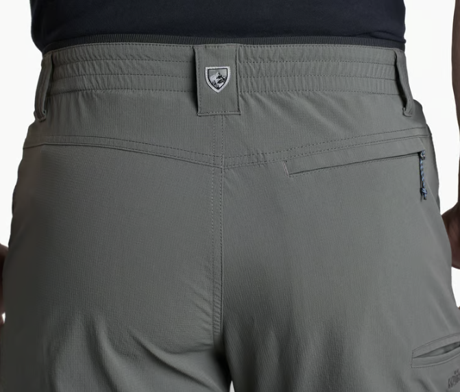 Close-up back view of a person wearing gray technical outdoor shorts, showing the waistband, belt loop, zip pocket, and a small shield-shaped logo patch.