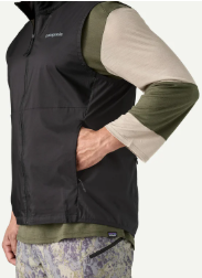 A close-up of a person wearing a black Patagonia zip-front vest over a long-sleeve shirt with olive green and beige sleeves, paired with patterned pants, against a light gray background.