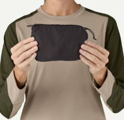 A person holding a small black drawstring pouch in front of their chest, wearing a beige and dark green long-sleeve shirt against a plain white background.